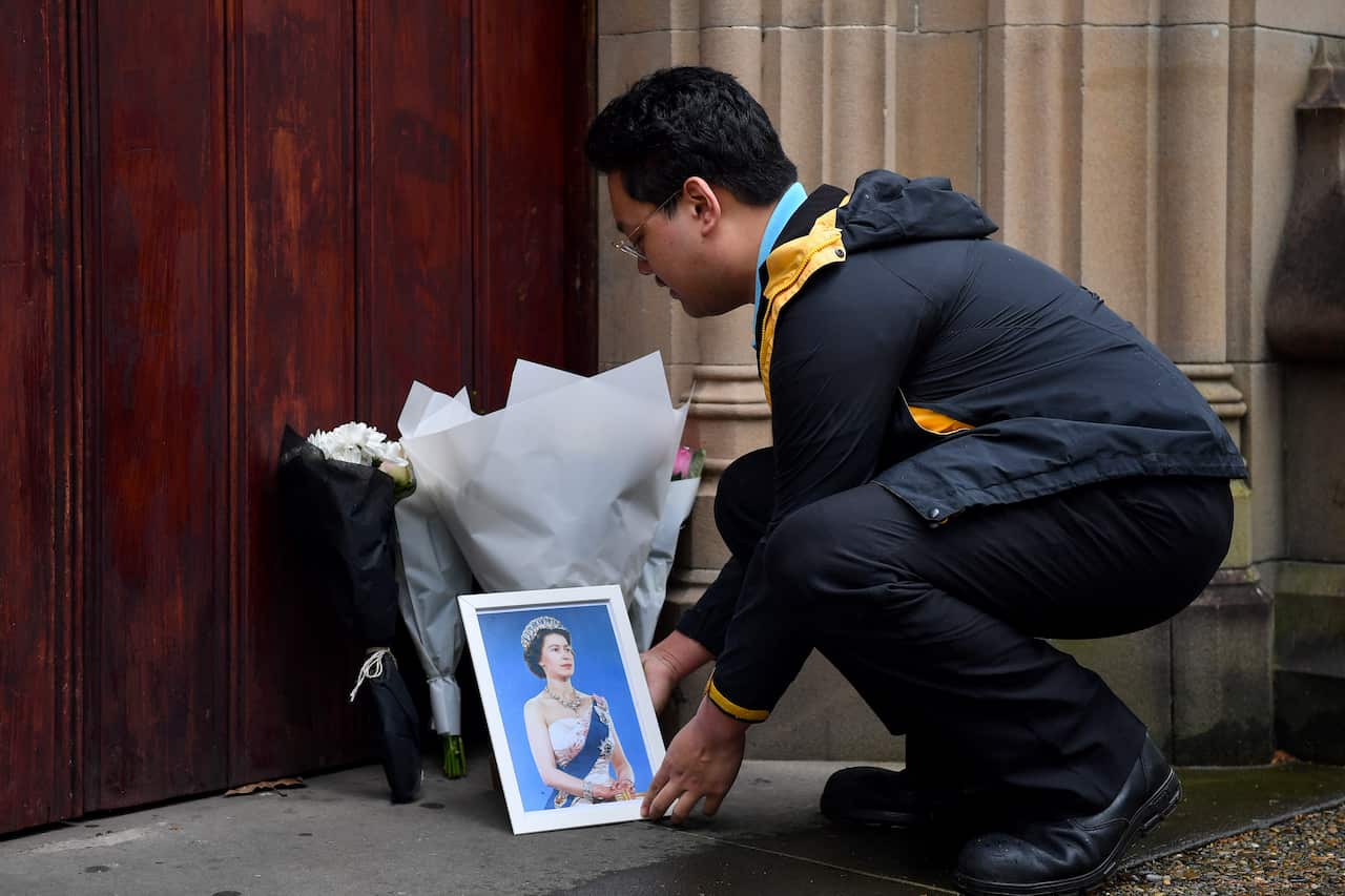 A man crouches in front of a portrait of Queen Elizabeth II and a few bunches of flowers.