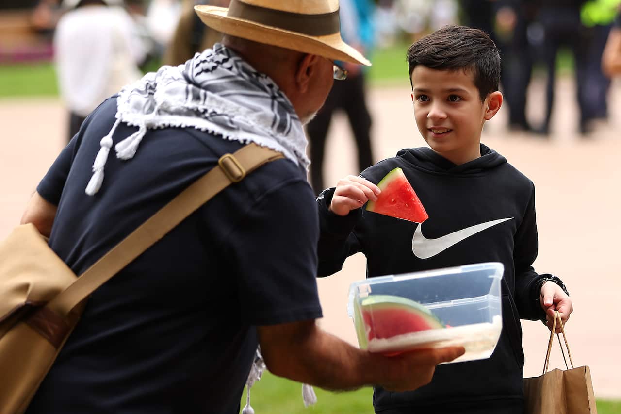 A man wearing a hat is offering watermelon pieces to a young boy.