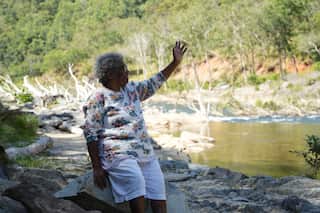 An older woman in a print-patterned white shirt and matching shorts sits on a rock by a lake, one hand raised.