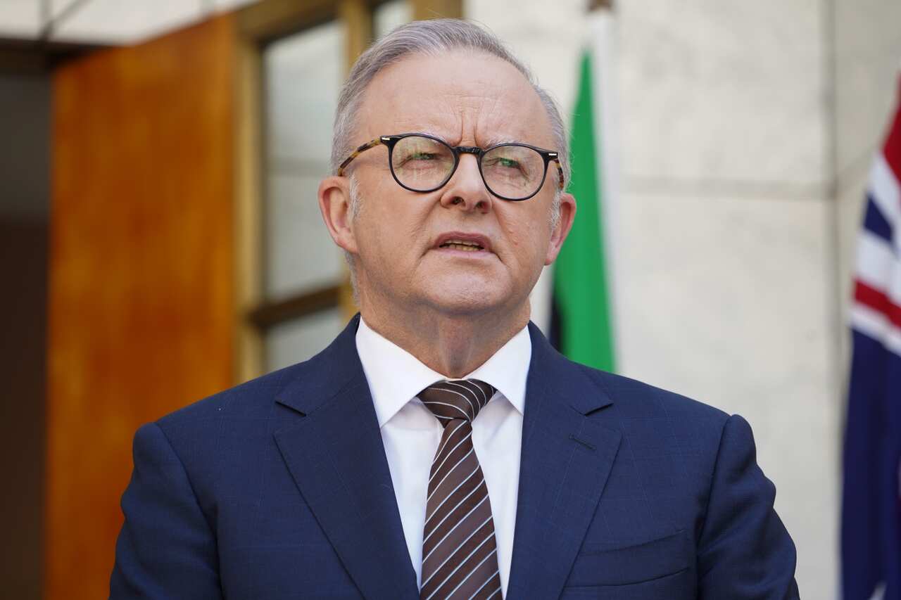 Anthony Albanese, wearing a blue suit, whit eshirt and brown tie, standing in front of a grey building.