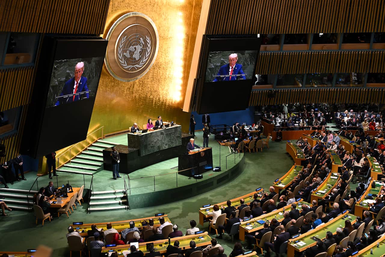 An aerial view of a large indoor chamber with a raised platform. Donald Trump is speaking from behind a lectern as people seated at curved benches listen