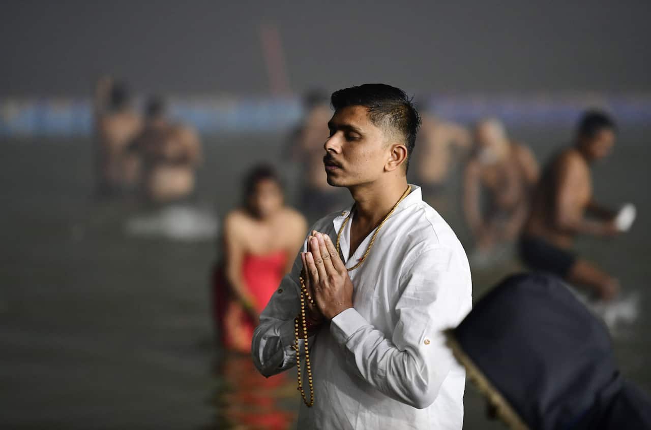 A moustachioed man in a white shirt holds prayer beads between his hands as he stands in the waters of a river.