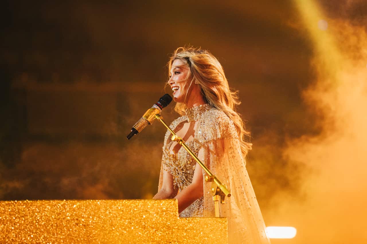 Delta Goodrem, wearing a gold-coloured gown, sits at a sparkly gold piano and sings into a microphone