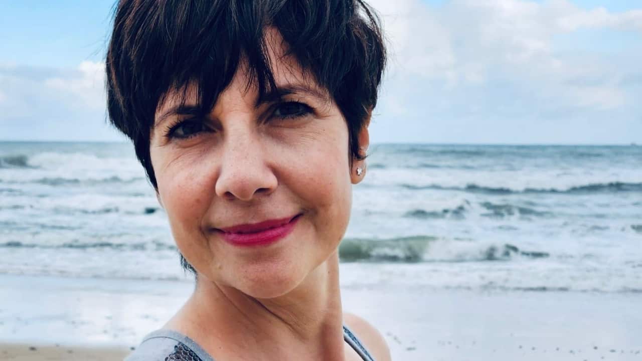 A close-up of a middle-aged woman with a dark pixie cut smiling while standing on the beach with waves in the background 