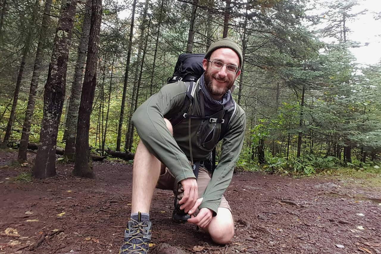 A white man wearing outdoor gear and a backpack kneels in some woods.