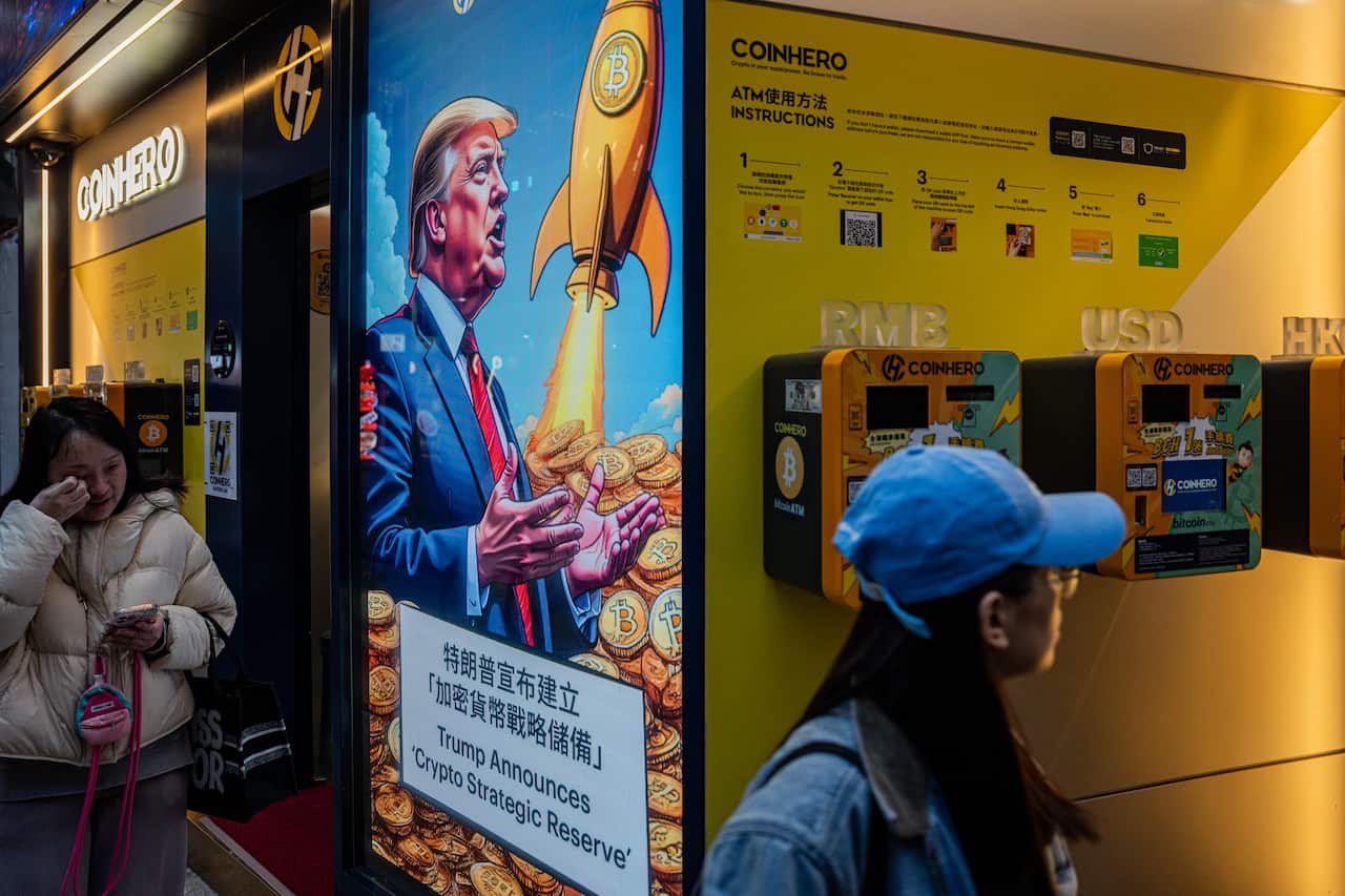 People walk past a screen showing Donald Trump speaking while surrounded by gold coins embossed with cryptocurrency logos and a rocket launching.