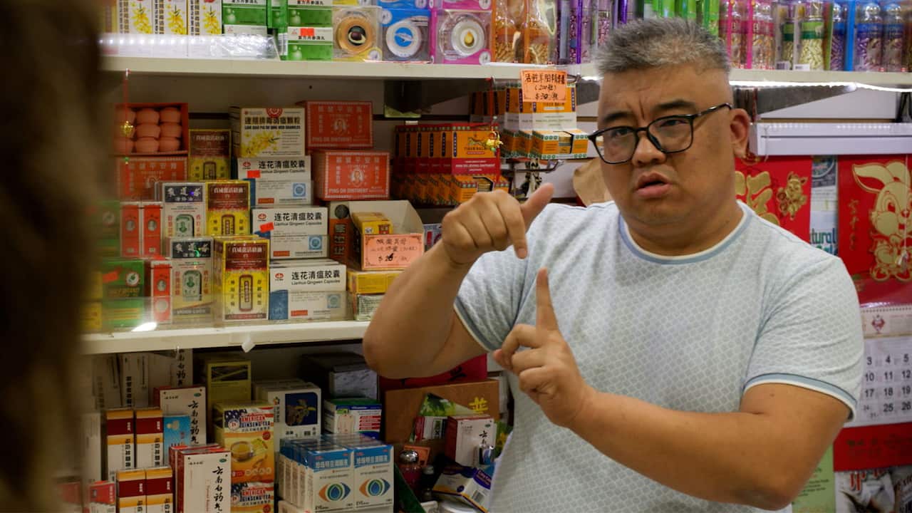 A middle-aged Asian man with short grey hair, wearing a grey t-shirt and reading glasses, is gesturing in sign language. He is standing at what appears to be a grocery store with shelves filled with goods in the background. 