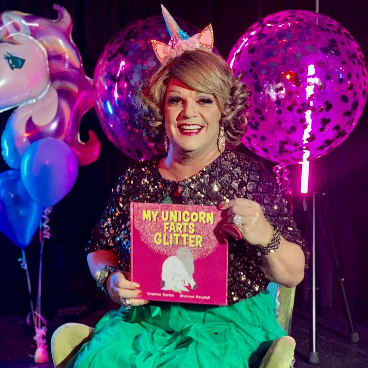 A drag queen in a green dress, glittery jacket, wearing a unicorn headband sit son a chair, holding a book called My Unicorn Farts Glitter. In the background are colourful balloons, one of which is in the shape of a unicorn's head
