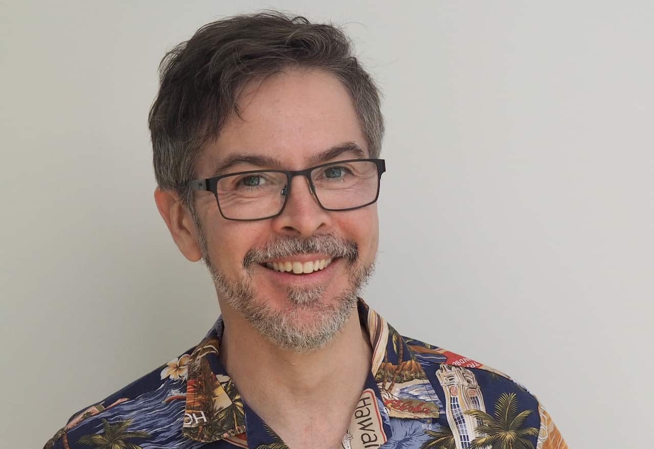 A man with grey hair and facial hair and glasses, smiling. He's wearing a tropical shirt.