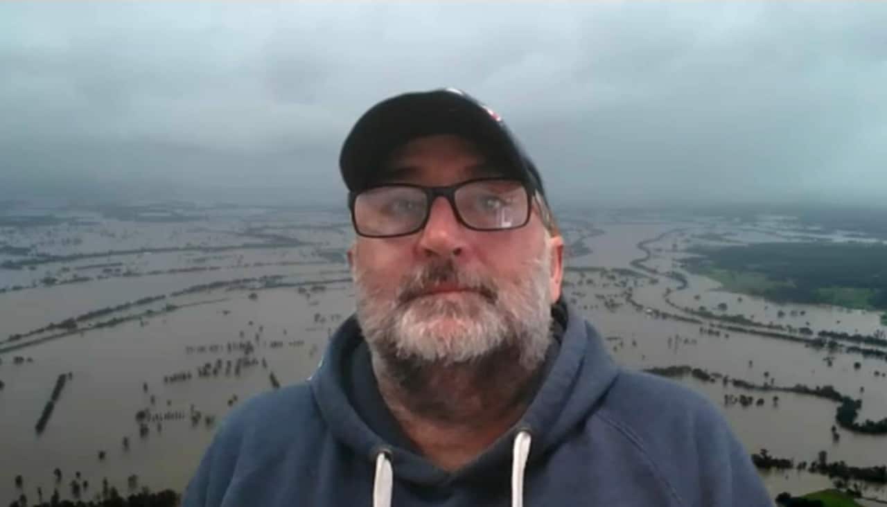 A screenshot of a zoom call, showing the face of a middle aged man with white beard and wearing a black cap, superimposed on a background of an aerial view of flooded farmland.