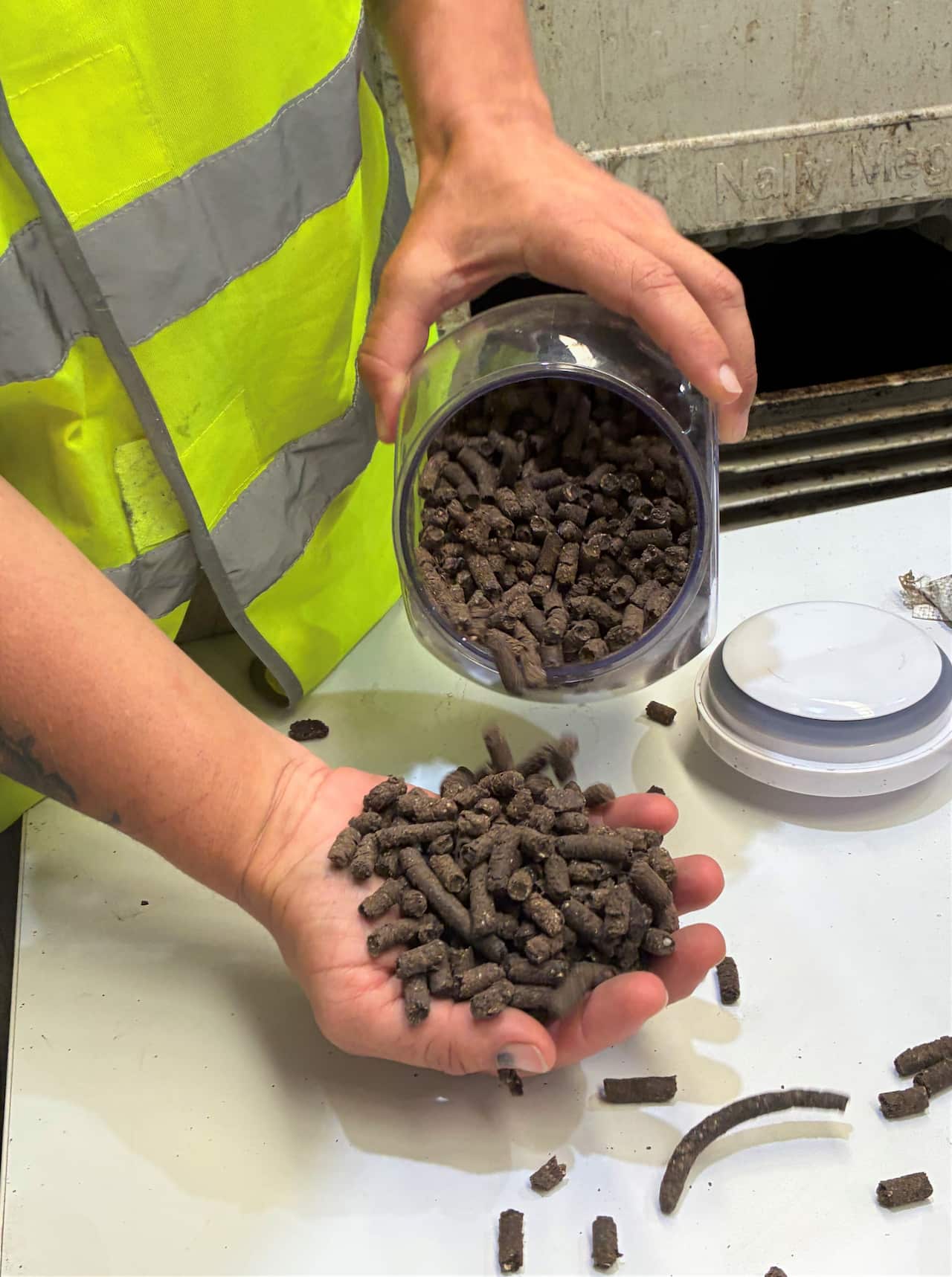 A hand holds brown pellets of fertiliser being poured from a glass jar.