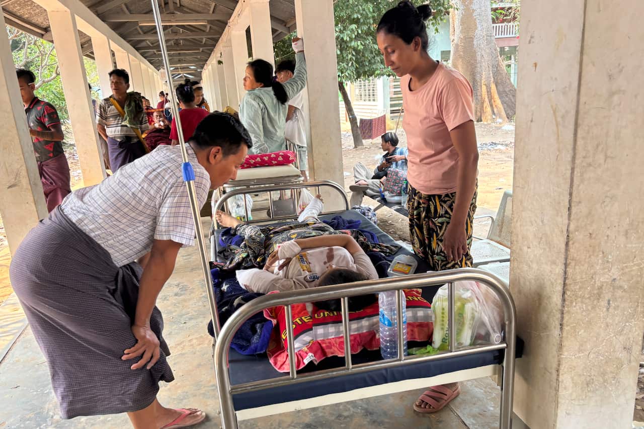 An injured person lying in a hospital bed, with two people standing over them. Others are in the background.
