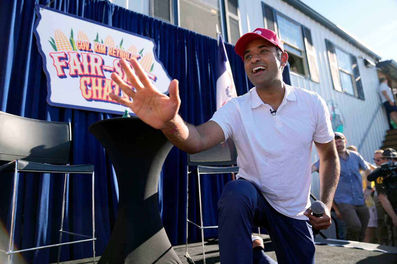 Vivek Ramaswamy wearing a light-coloured polo shirt and red cap and kneeling on a stage