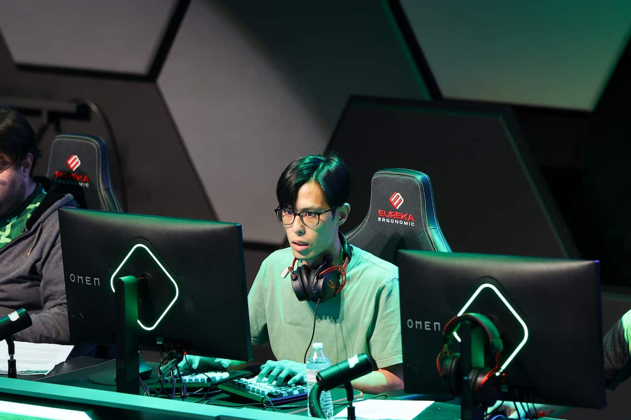 A young man is sitting at a computer with headphones around his neck. 