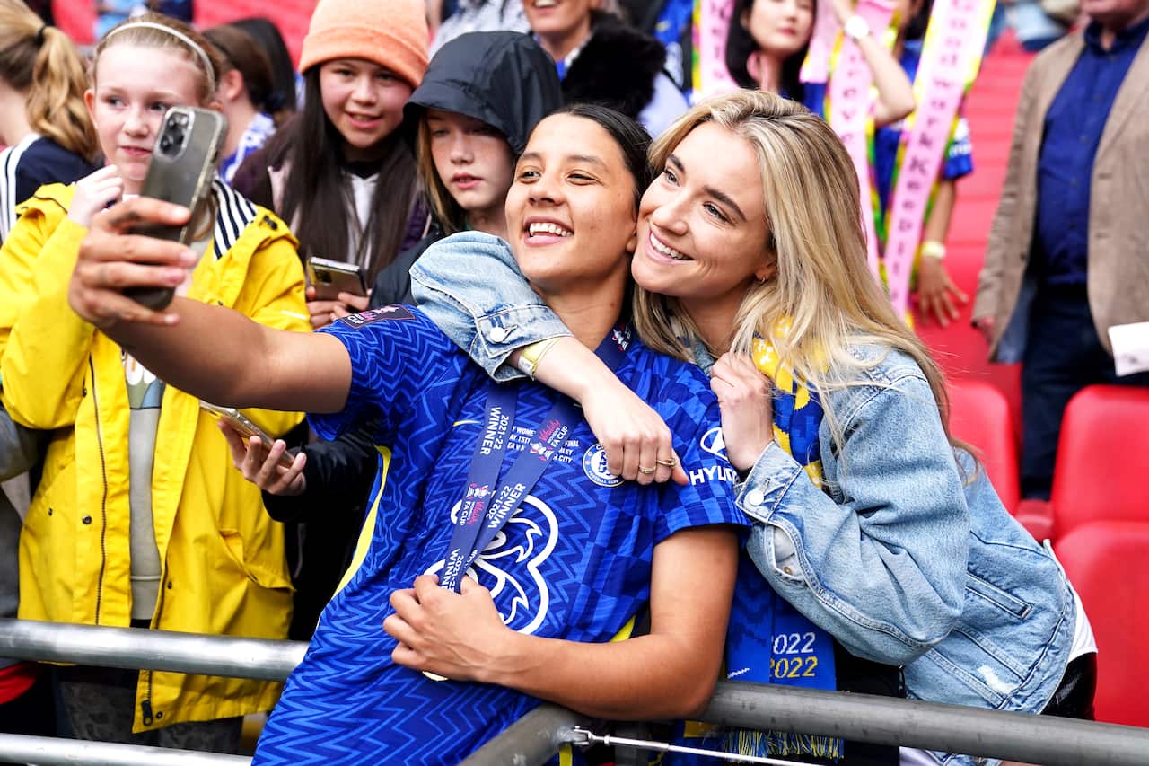 A woman footballer wearing a medal poses for a selfie with a woman in the stands