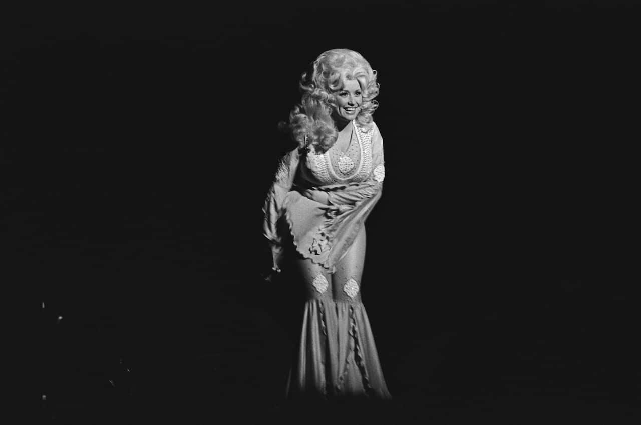 Black-and-white stage photo of a performer in a long embellished gown, standing alone under a spotlight against a dark background.