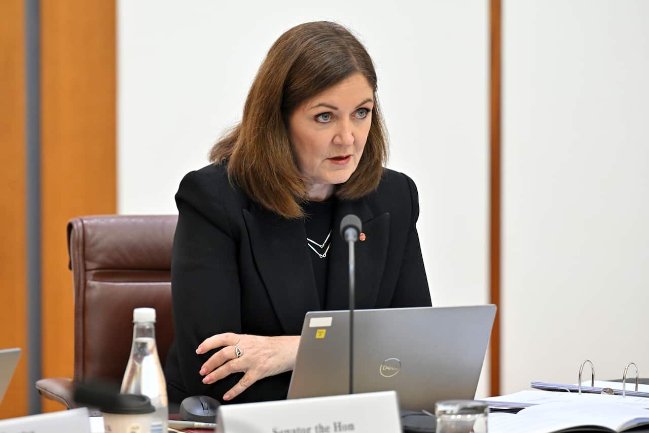 A woman wearing a black jacket and top sits at a desk with a grey laptop in front of her.