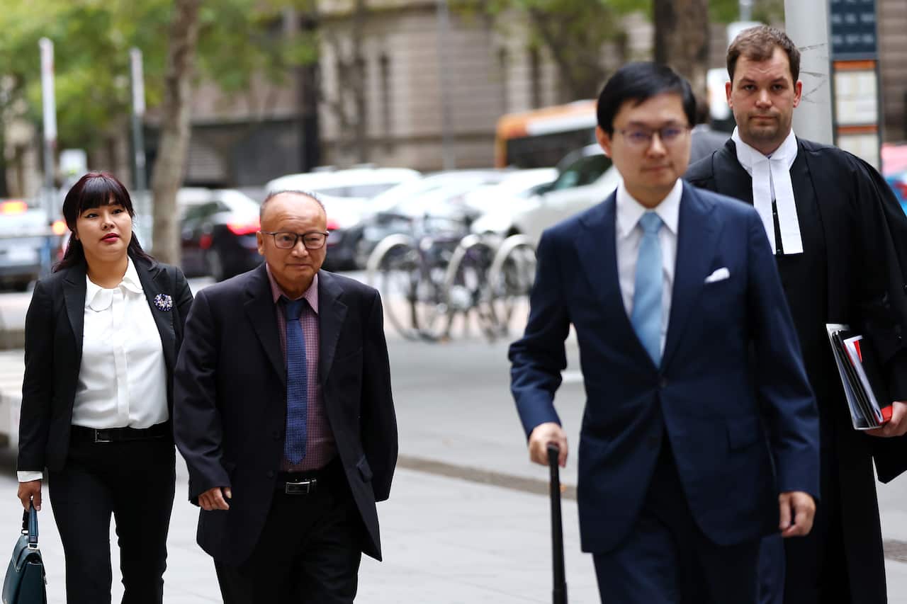 An elderly man wearing a suit walks along a footpath flanked by several others. One is wearing a Judge's gown.