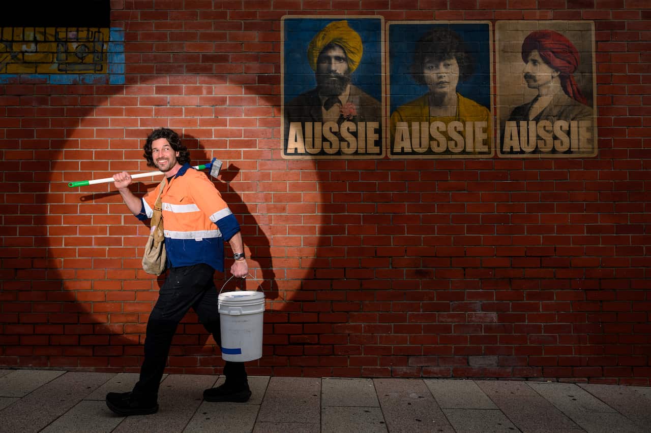 A man wearing a fluorescent orange shirt holds a broom, bucket and bag with a spotlight shining on him. On the brick wall behind him, there a posters of people and the text 'AUSSIE'.