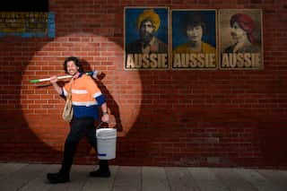 A man wearing a fluorescent orange shirt holds a broom, bucket and bag with a spotlight shining on him. On the brick wall behind him, there a posters of people and the text 'AUSSIE'.