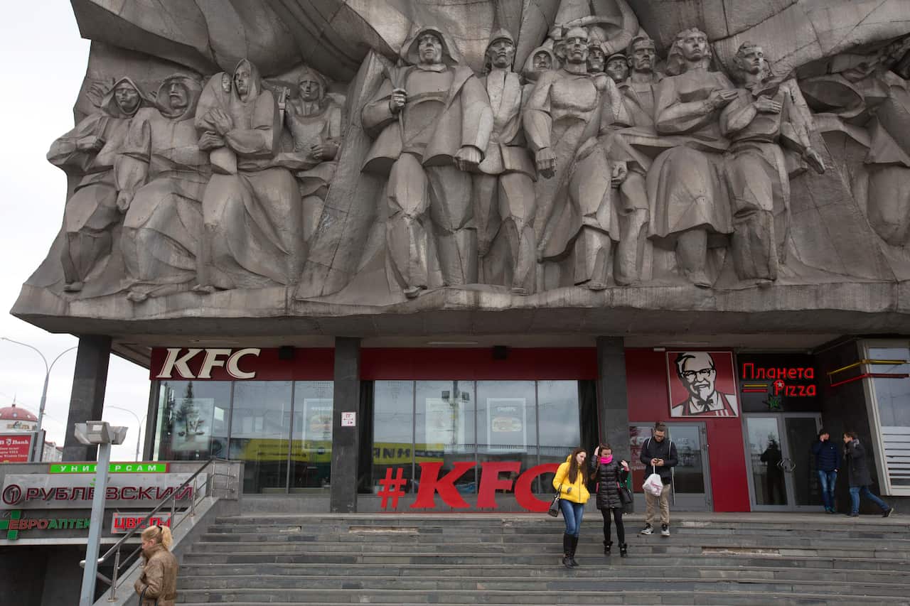 A large, brutalist concrete bas-relief monument featuring a group of figures looms over a ground floor KFC restaurant, with people walking on a set of steps in the foreground.