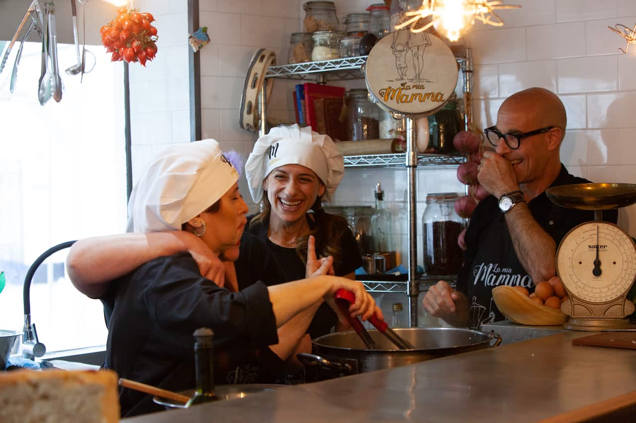 A man and two laughing women stand in a kitchen. Oneof the woman reaches into a large pot with a pair of red-handled tongs.