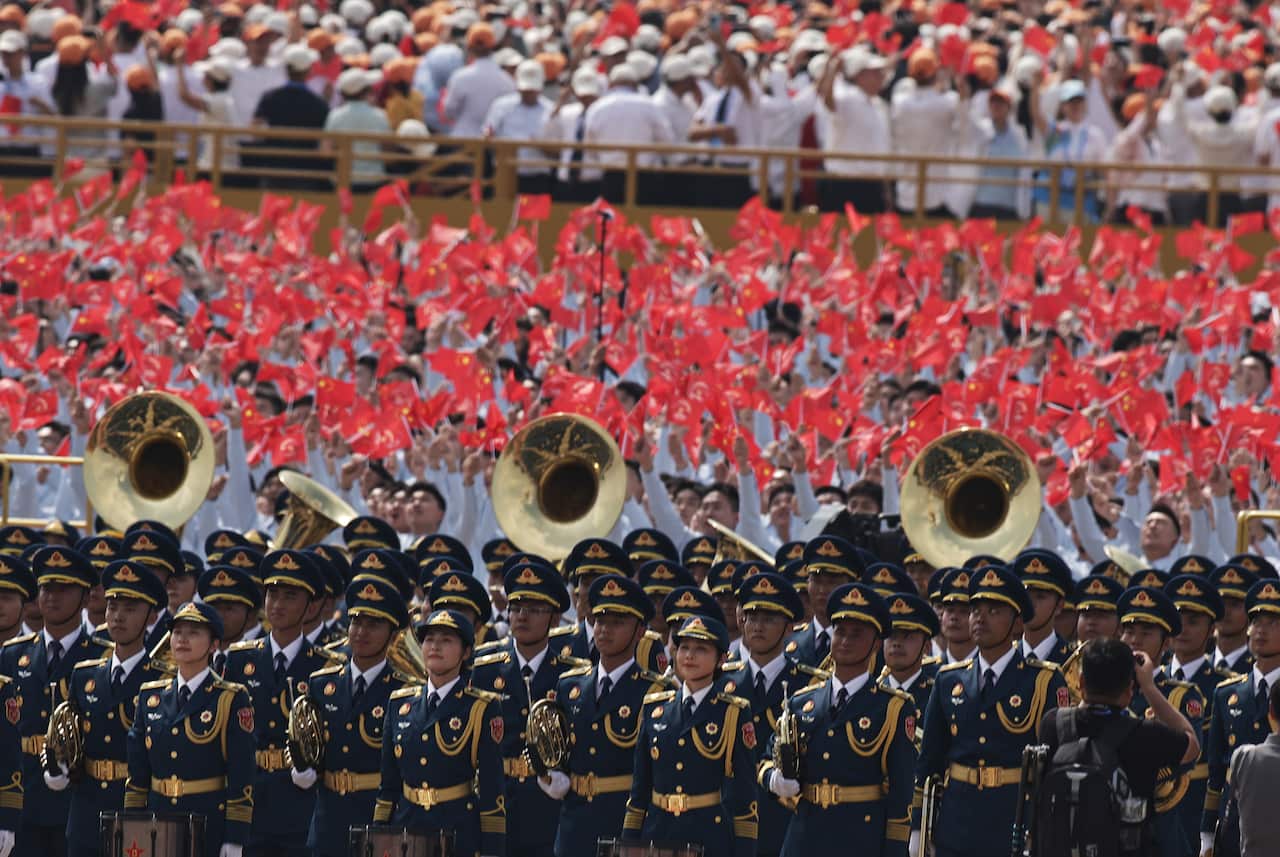 A Chinese military band performs during a military parade. There are rows of personnel holding red flags behind the band.