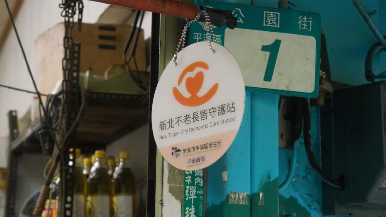 A round sign hanging outside a storefront, with Chinese text on it and the English text reading "New Taipei City Dementia Care Station"