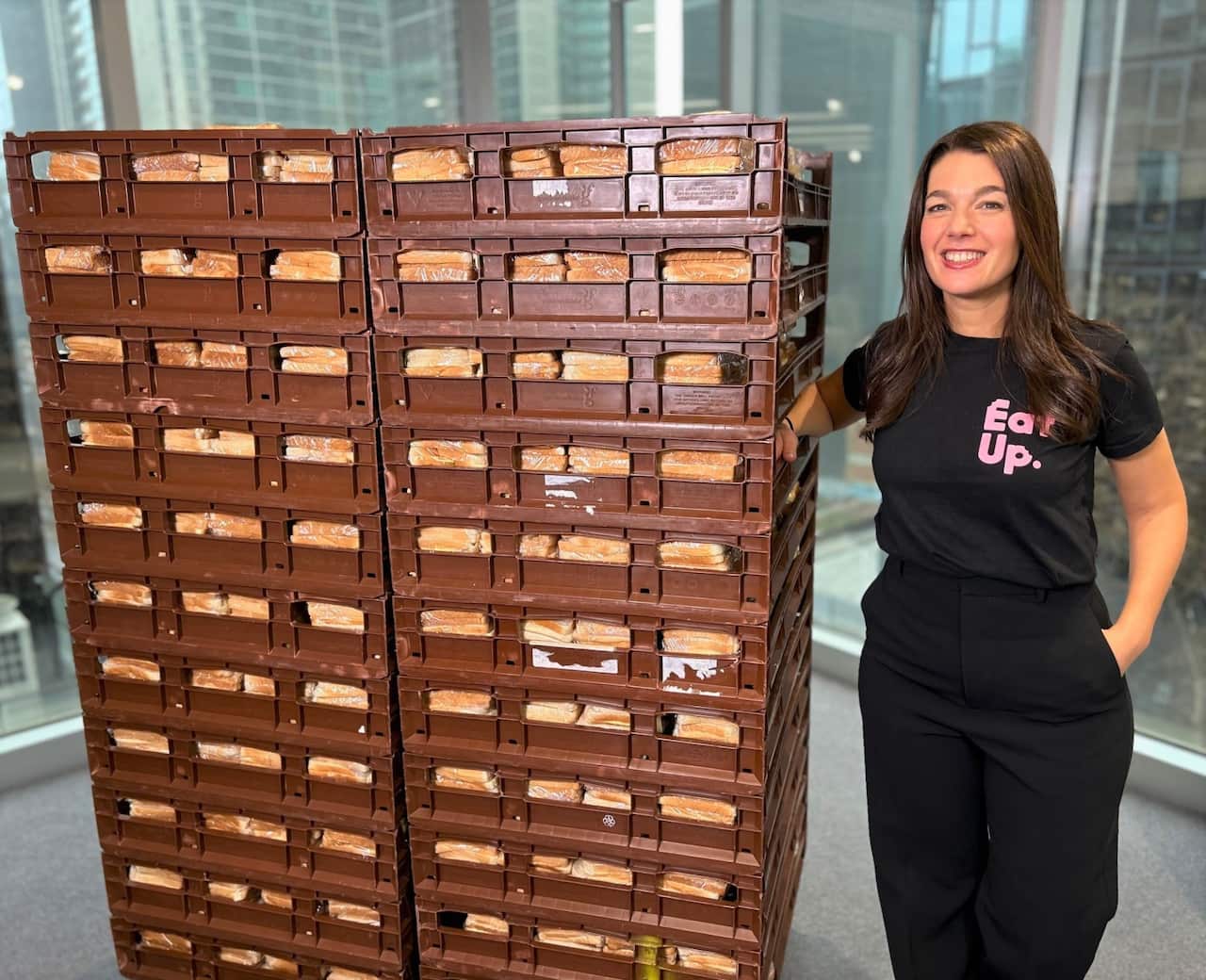 A woman in a black t-shirt stands next to a tall tower of sandwiches in crates.