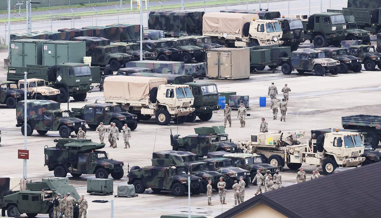 Military troops in tan and black vehicles on concrete seen from a distance.