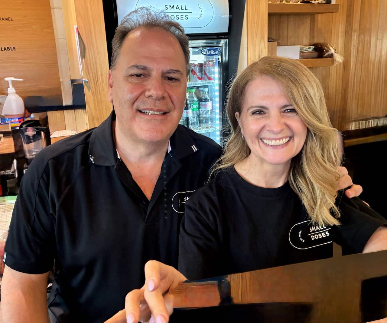 A man in a black t-shirt stands next to a woman with long blonde hair by the till of a cafe.