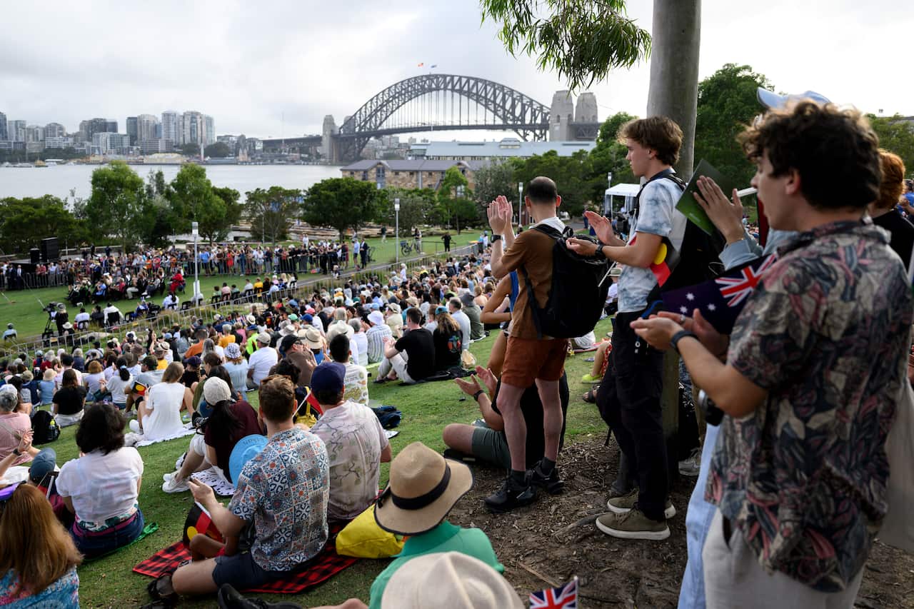 A large group of people sitting in a park. The Sydney Harbour Bridge and skyline are in the distance.