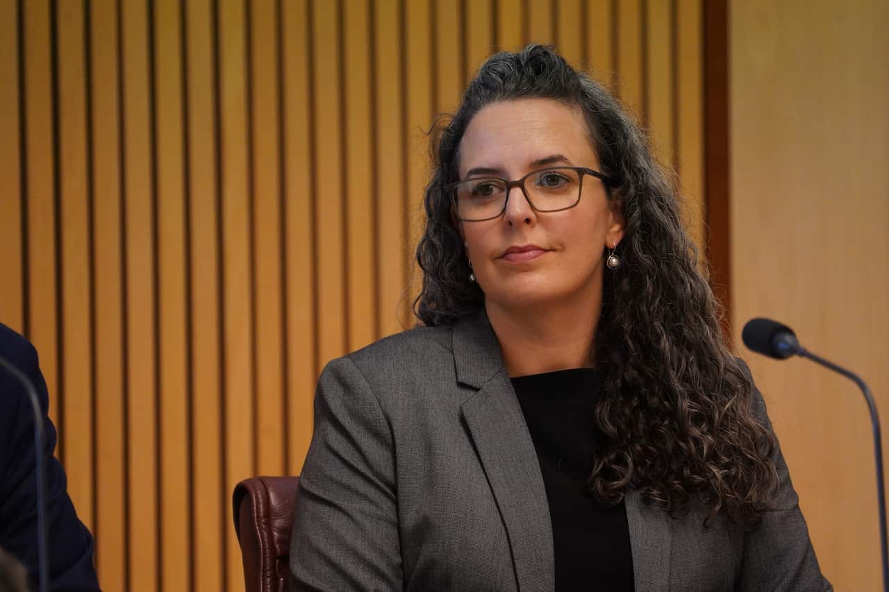 A woman with long curly hair, wearing a grey blazer, sits in front of a microphone, looking sideways.