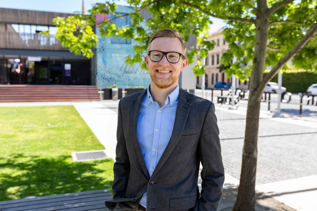 A man in a dark suit worn over a blue shirt smiles as he stands outside next to a tree.