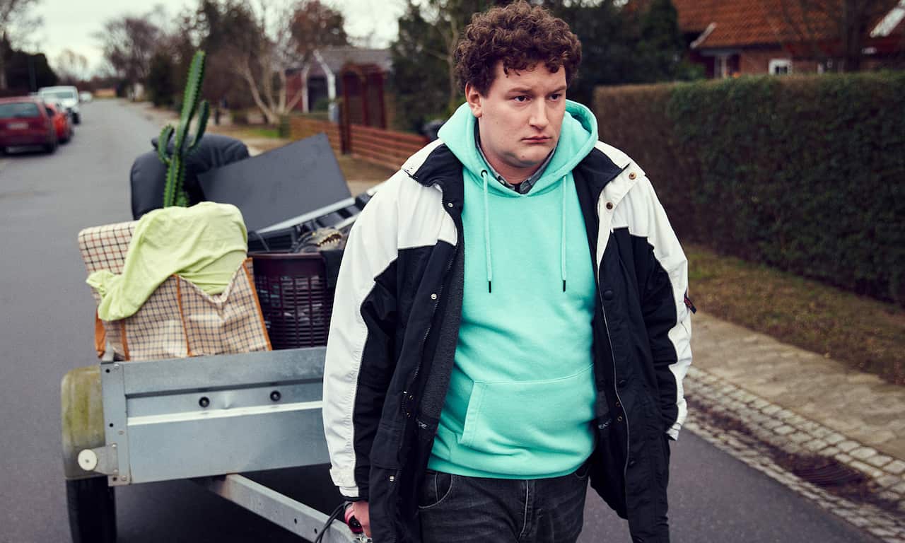 A young man in mint green hoodie and black and white jacket drags a trailer along a street. A computer monitor can be seen among the bags and baskets in the trailer.