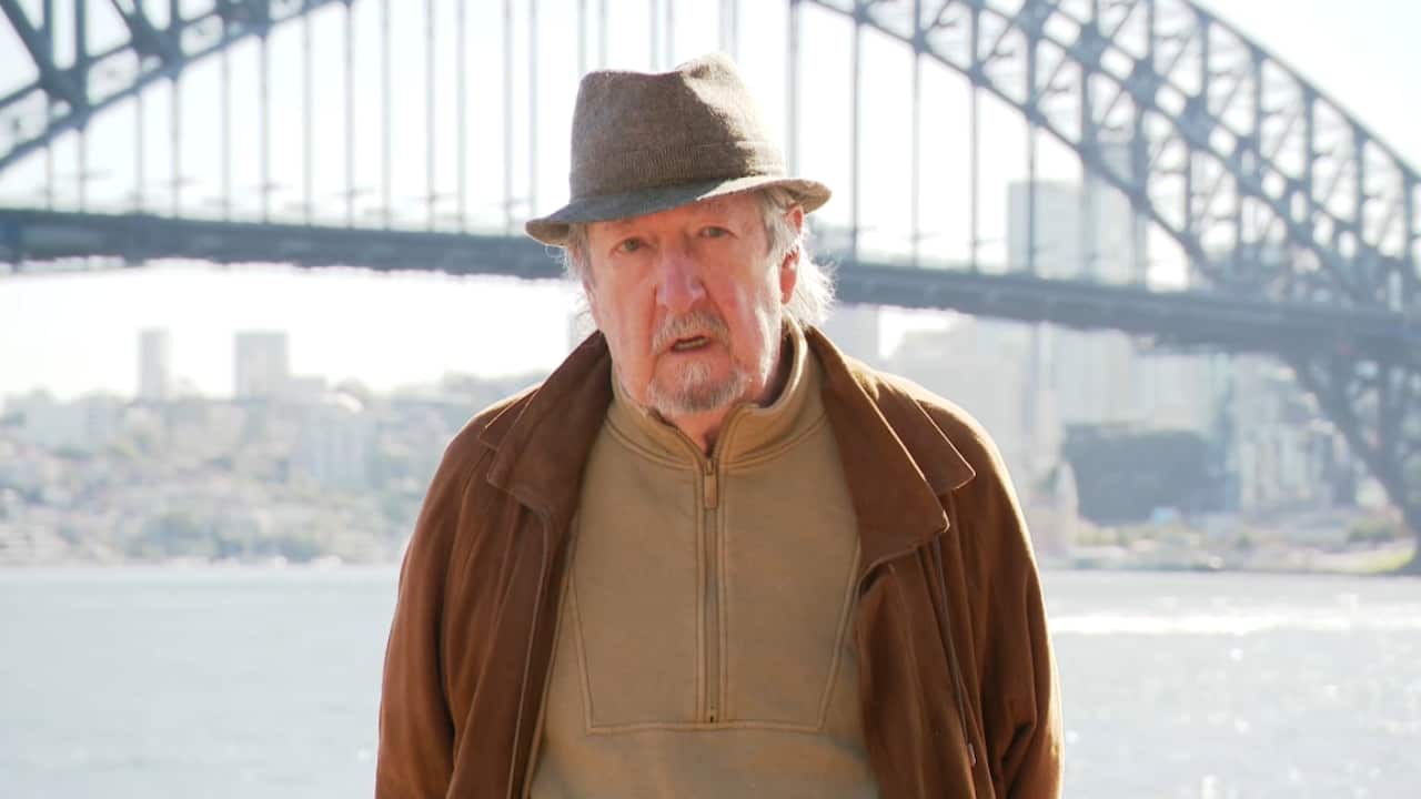 An old white man in a tan jumper, brown jacket and grey fedora stands in front of the Syndey Harbour Bridge