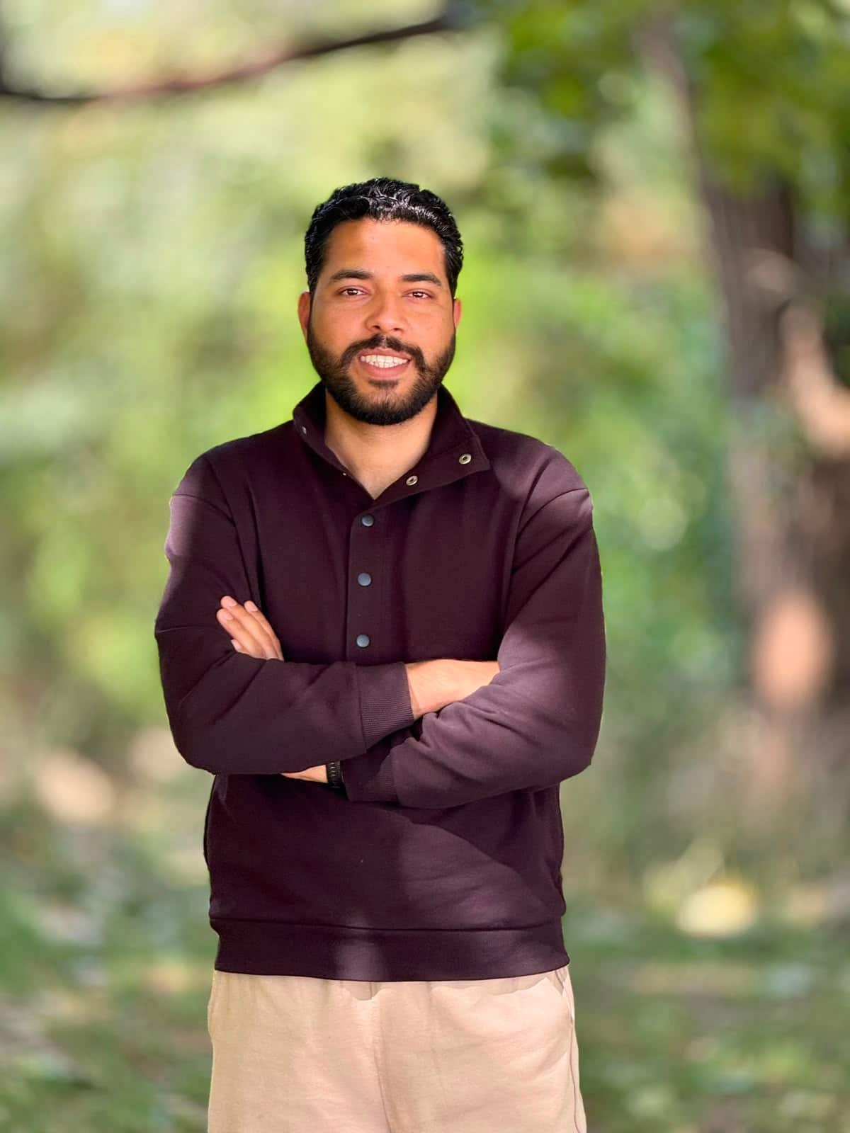 A man in a black shirt stands in a garden, his arms folded.