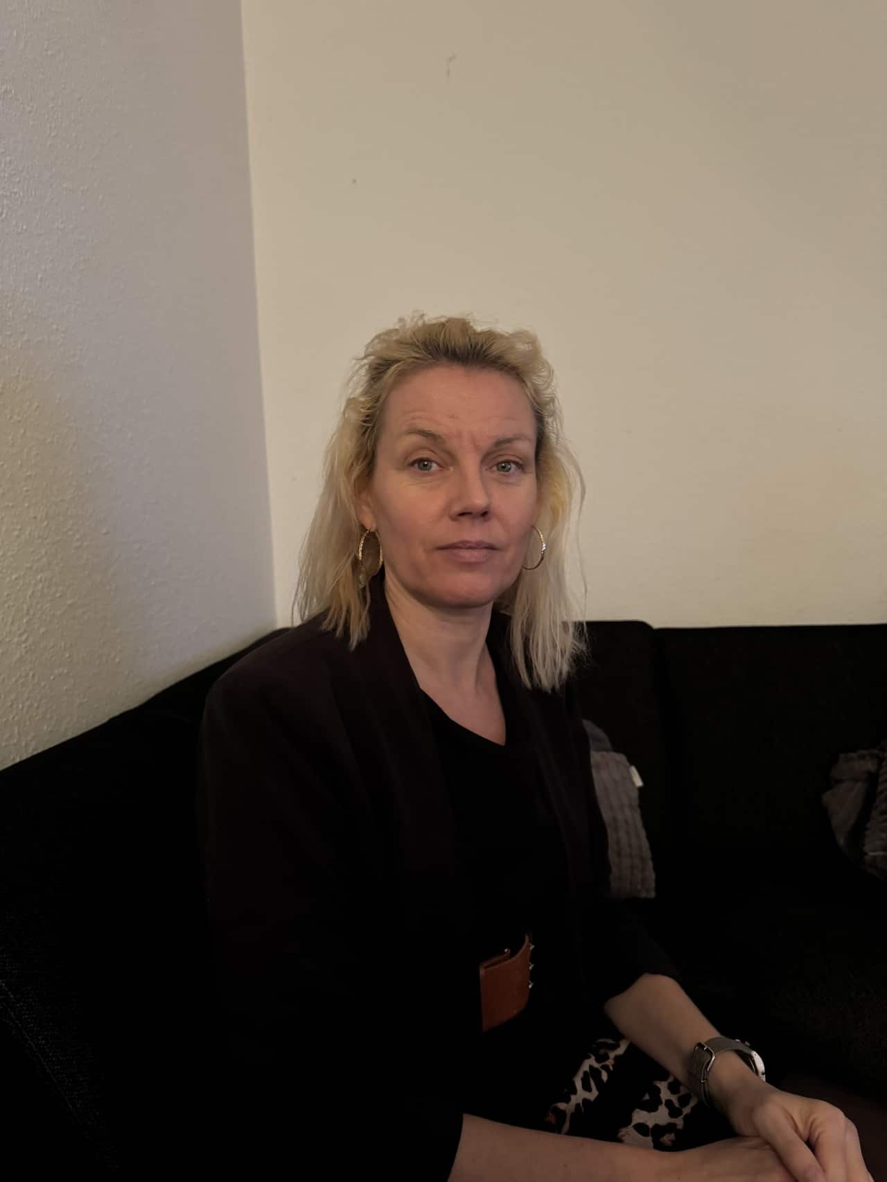 A blonde woman with silver hoop earrings sits on a couch facing the camera. She has a neutral facial expression. 