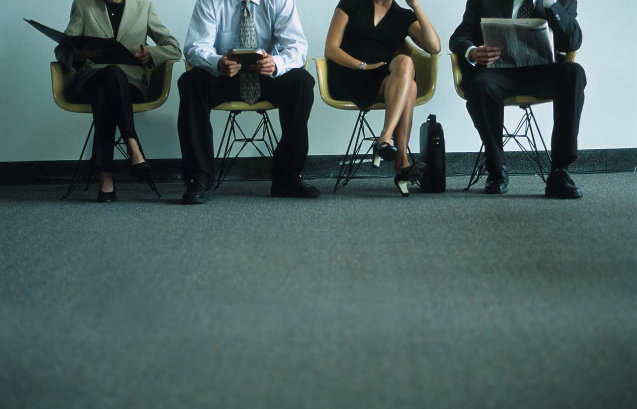 Office workers sitting in chairs lined up against a wall.