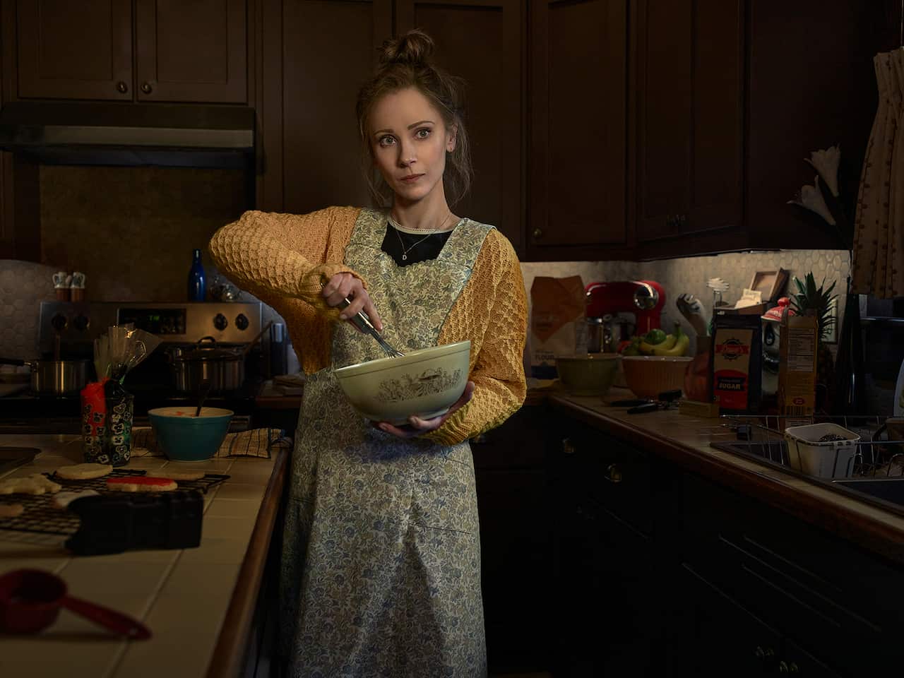Juno Temple stands in a dimly lit kitchen wearing and apron and stirring the contents of a bowl that she holds in front of her.