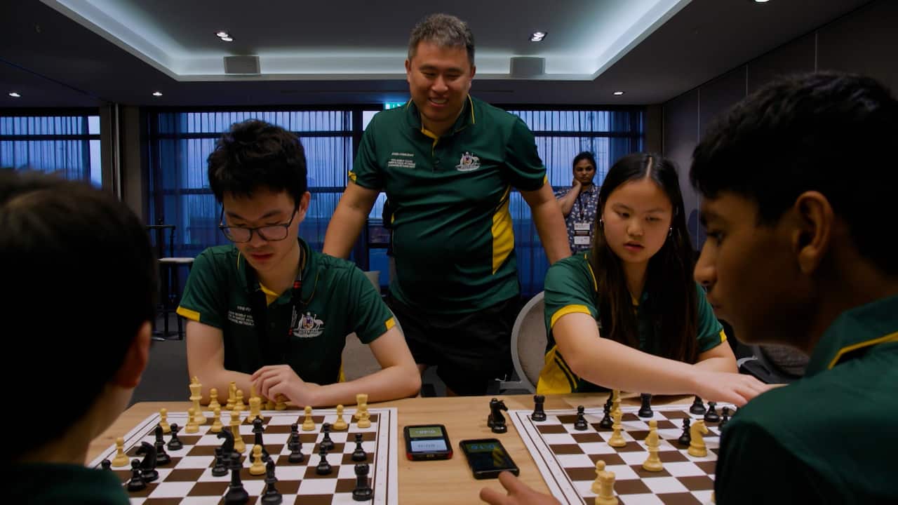 A man stands behind a teenage boy and girl playing chess. They all wear green-and-yellow polos representing the Australian national team