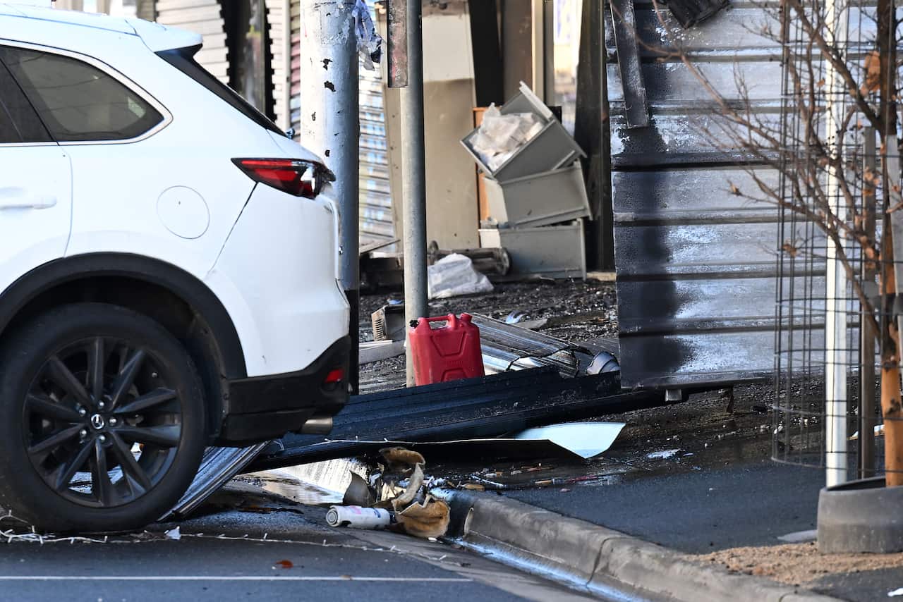 A white SUV parked next to a storefront showing signs of fire damage, with debris, a red jerrycan, and charred building materials scattered on the ground.