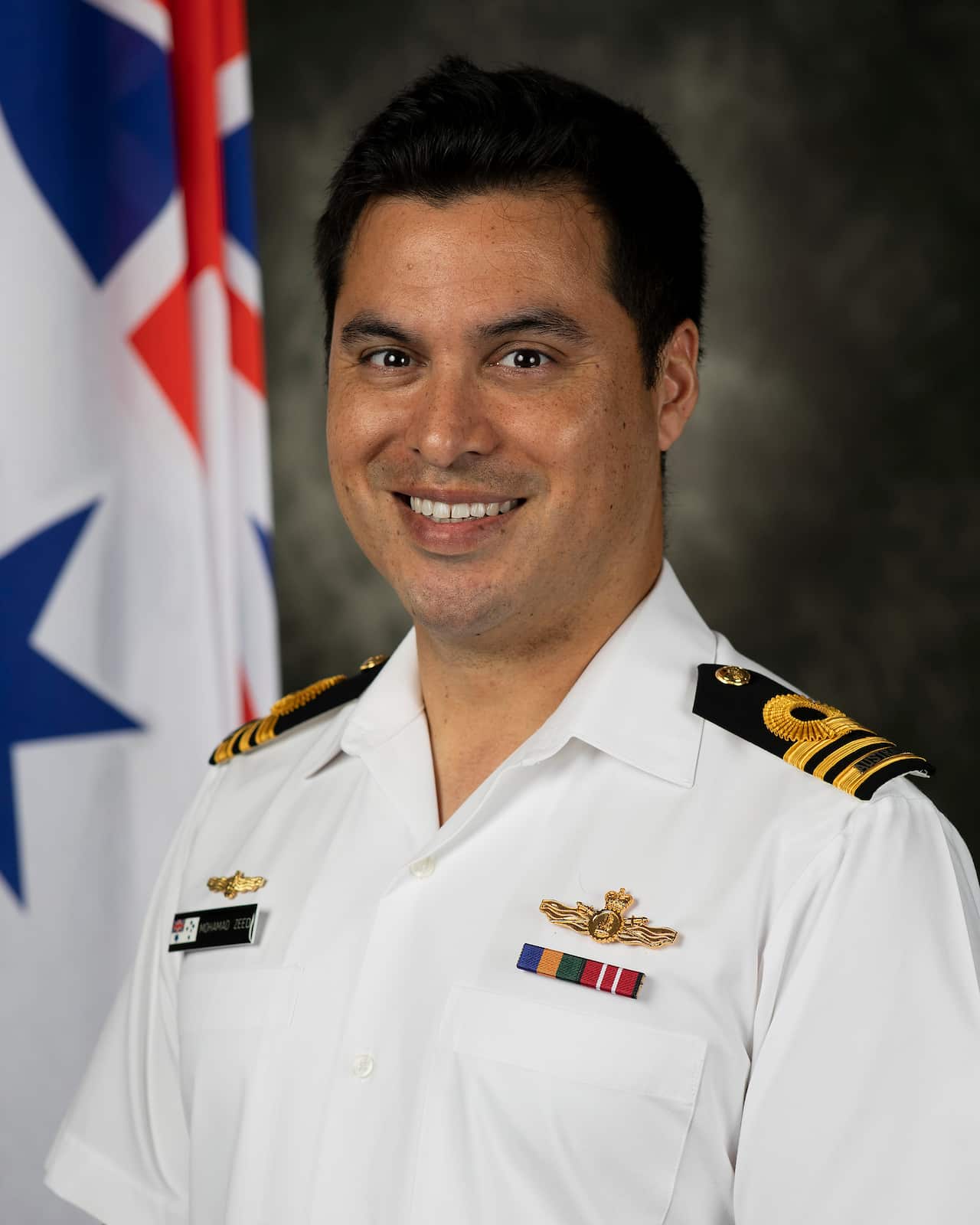 A medium shot of a smiling man in a white Royal Australian Navy officer uniform standing in front of an Australian White Ensign flag.