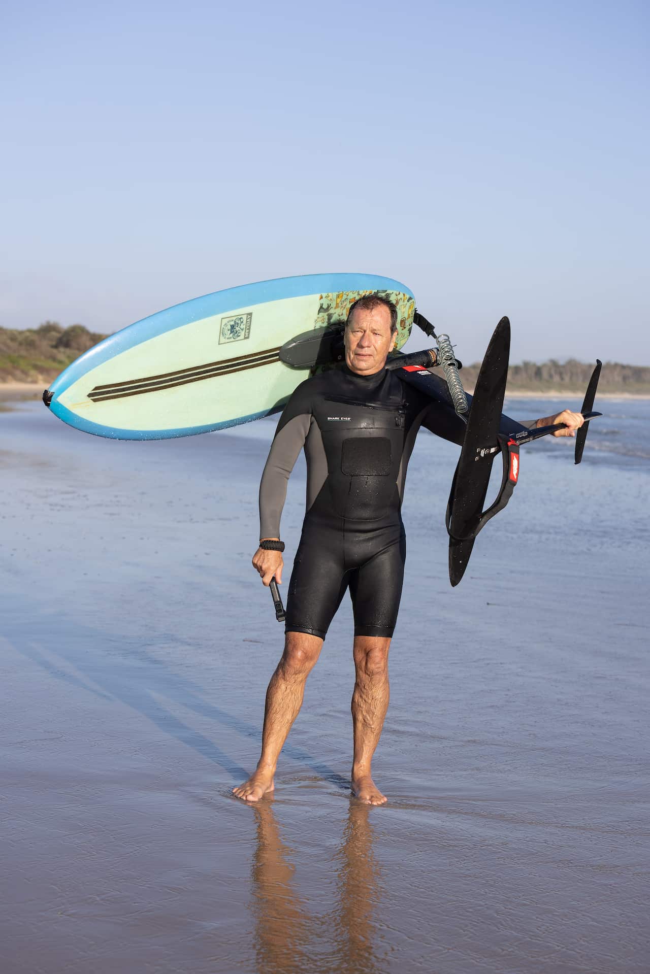 A man in a wetsuit carries a surfboard with a hydrofoil attached.