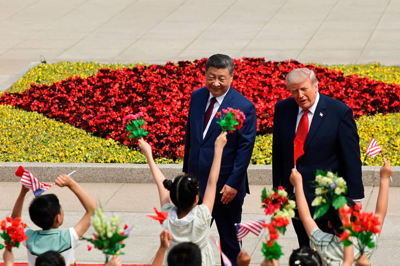 Two men in suits smile at a group of school children who have their hands in their air and hold flowers.