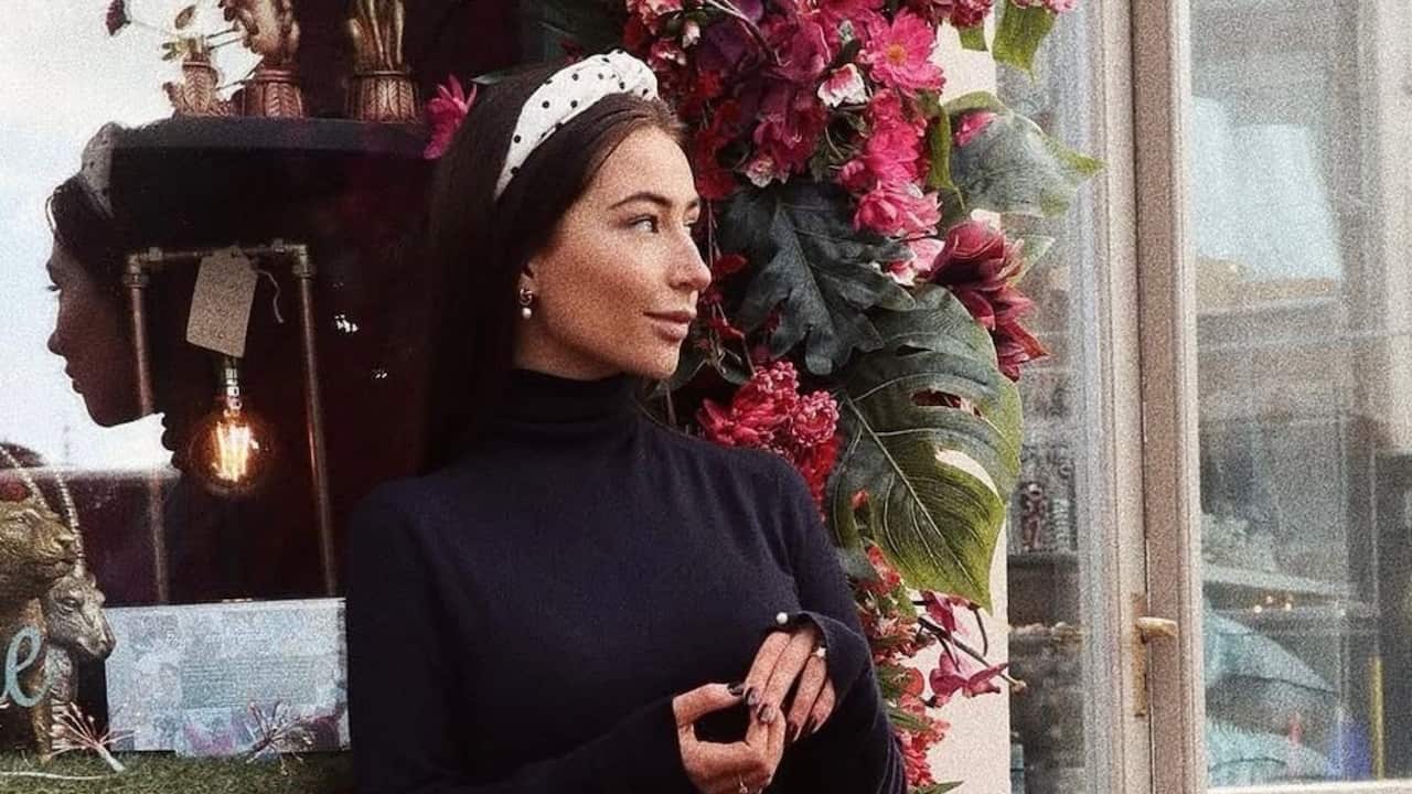 a young woman in a black dress and white headband looks off to the camera and sits in front of a shop window with flowers and fronds in the background