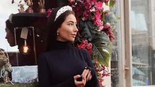 a young woman in a black dress and white headband looks off to the camera and sits in front of a shop window with flowers and fronds in the background