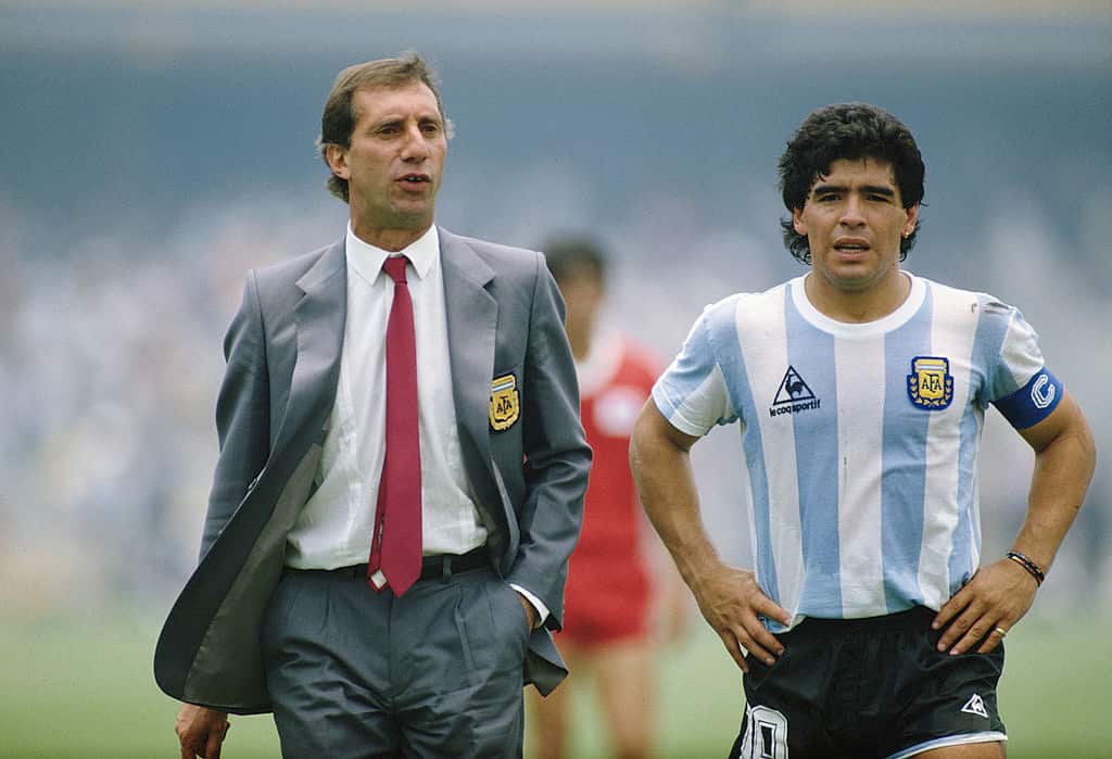 A man in a grey suit and red tie standing next to Diego Maradona, who is wearing a light blue and white striped football shirt and black shorts
