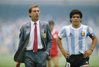 A man in a grey suit and red tie standing next to Diego Maradona, who is wearing a light blue and white striped football shirt and black shorts