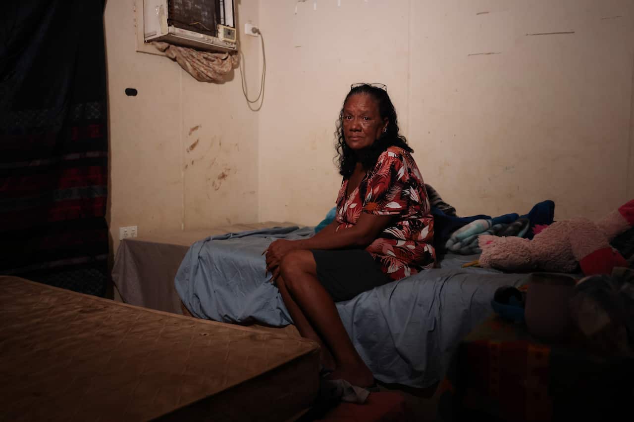 A middle-aged woman looking into the camera lens, sitting on a mattress, with an air conditioning machine in the background.